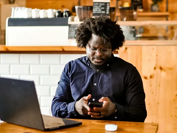 Focused man working remotely at coffee shop using smartphone and laptop on wooden table surrounded by warm decor